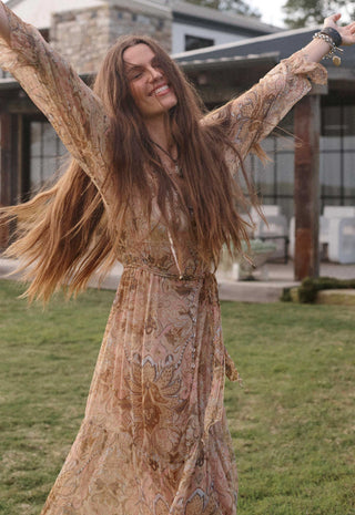 A woman with long brown hair wearing a flowing beige and brown patterned bohemian maxi dress, posing outdoors with her arms raised in front of a modern house and grassy lawn.