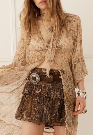 A woman wearing a bohemian-style outfit featuring a sheer beige paisley cardigan, a brown patterned ruffled mini skirt, and a wide brown belt with a silver buckle.