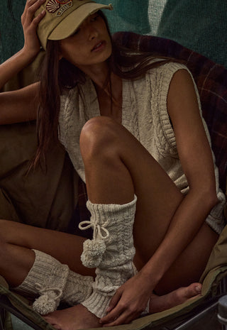 a-woman-seated-in-a-camp-chair-wearing-a-beige-knit-sleeveless-top-matching-knit-leg-warmers-with-pom-poms-a-tan-baseball-cap-and-olive-pants-in-a-rustic-outdoor-setting