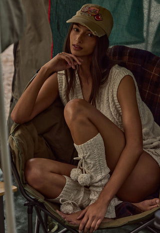 a-woman-sits-in-a-camping-chair-wearing-a-beige-knit-sleeveless-top-matching-knit-leg-warmers-and-a-beige-baseball-cap-with-a-red-embroidered-bee