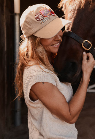 a-woman-in-a-cream-knit-top-and-beige-baseball-cap-with-a-horse-smiling-in-a-barn-setting-the-cap-features-a-colorful-embroidered-logo-on-the-front