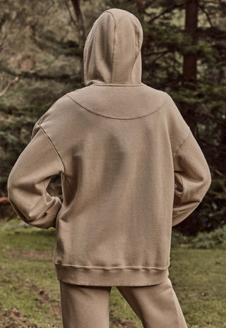 A person viewed from behind wearing a matching beige hooded sweatshirt and jogger pants in an outdoor wooded setting.