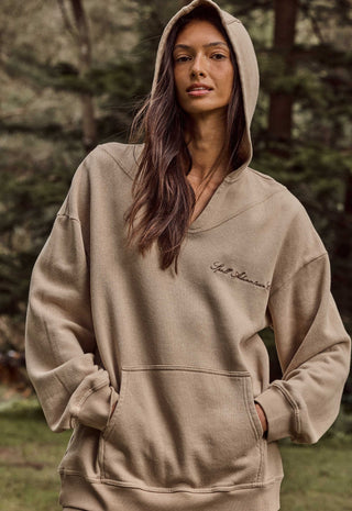 A woman wearing a beige oversized pullover hoodie with a small script logo on the left chest, photographed outdoors against a blurred forest background.