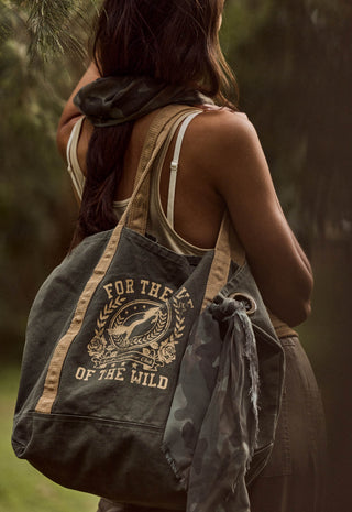 a-woman-outdoors-carrying-a-large-canvas-tote-bag-over-her-shoulder-the-bag-is-dark-olive-green-with-tan-straps-and-a-tan-graphic-print-that-reads-for-the-love-of-the-wild-around-a-deer-illustration