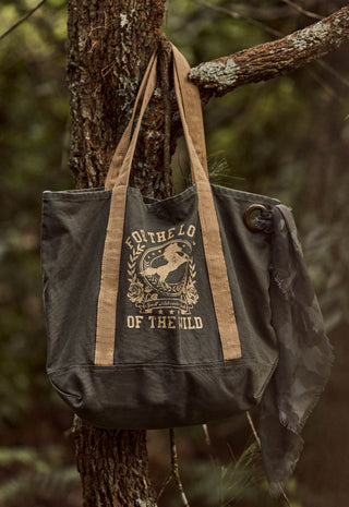 a-dark-gray-canvas-tote-bag-with-tan-straps-and-a-distressed-cream-graphic-reading-for-the-love-of-the-wild-hanging-from-a-tree-branch-in-a-forest-setting