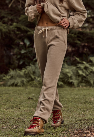 a-person-outdoors-wearing-loose-beige-jogger-sweatpants-with-a-drawstring-waist-a-matching-beige-cropped-sweatshirt-and-brown-hiking-boots-with-red-laces-standing-on-grass-with-a-blurred-forest-background