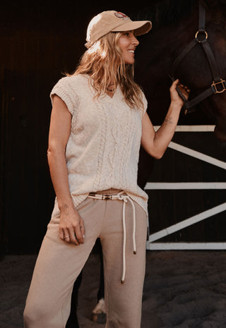 a-woman-wearing-a-beige-knit-sleeveless-top-matching-beige-drawstring-pants-and-a-beige-baseball-cap-stands-in-a-barn-next-to-a-dark-horse-holding-its-bridle-reins