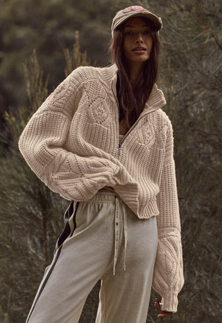 A woman wearing a chunky cable-knit zip-front sweater in a warm cream or oatmeal tone, paired with light beige sweatpants and a beige cap, standing outdoors against a blurred natural background.