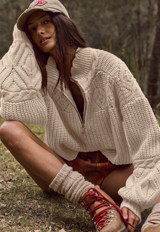 A woman crouching outdoors in a wooded setting, wearing a chunky cream cable-knit half-zip sweater, a matching beige baseball cap, brown plaid shorts, and tan lace-up boots with red laces.
