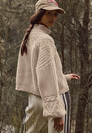 A woman wearing a chunky cream cable-knit mock-neck sweater, a beige baseball cap, and cream pants with black stripes, standing in a wooded outdoor setting.