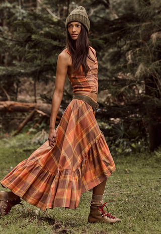 A woman wearing a rust-orange and brown plaid tiered maxi skirt with a matching cropped top, a green knit beanie, and brown lace-up boots, standing in a grassy forest clearing.
