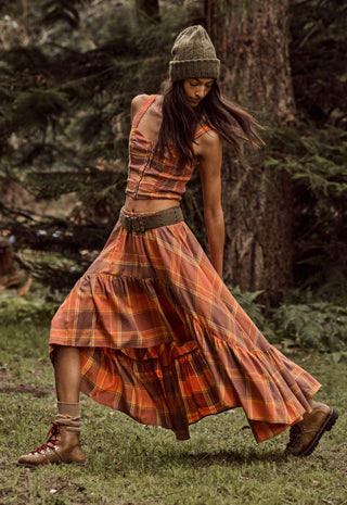 A woman wearing a rust-orange and brown plaid tiered maxi dress with a matching belt, a knit beanie, and lace-up boots, captured mid-motion while walking in a forest.