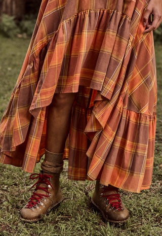 A close-up outdoor shot of a person wearing a long tiered plaid skirt in rust orange, brown, and yellow tones, paired with brown lace-up hiking boots on grass.