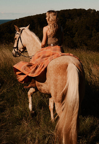 A woman riding a light-colored horse through a grassy outdoor landscape at golden hour, wearing a brown top and a flowing orange plaid skirt.