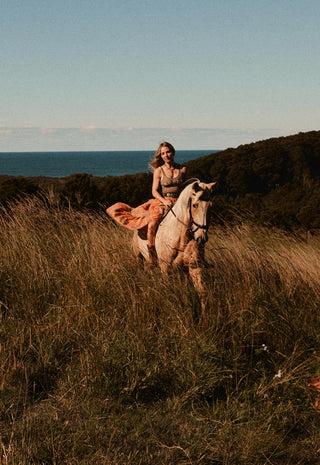 A woman rides a light-colored horse through tall dry grass on a hillside, with ocean and dark shrubbery in the background under a pale blue sky.