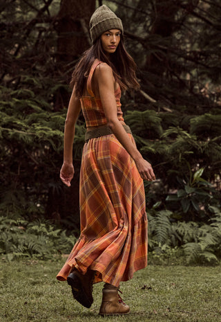 A woman wearing a sleeveless orange and brown plaid maxi dress with a matching beanie and brown boots, standing in a grassy outdoor setting with trees in the background.