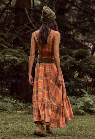 A woman viewed from behind wearing a sleeveless orange and brown plaid maxi dress with a matching belt and a knit beanie, standing on grass in a wooded outdoor setting.