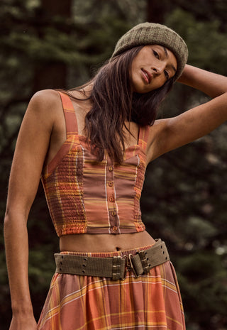 a-woman-wearing-a-rust-orange-and-brown-plaid-sleeveless-crop-top-with-button-details-a-matching-plaid-skirt-and-a-textured-olive-green-beanie-photographed-outdoors-against-a-blurred-forest-background