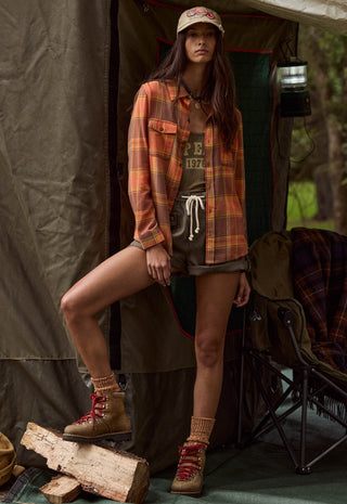 a-woman-posing-in-front-of-a-camping-tent-wearing-a-rust-orange-and-brown-plaid-flannel-shirt-over-a-graphic-tee-olive-shorts-and-hiking-boots