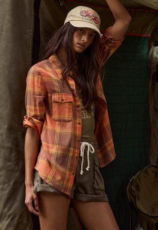 a-woman-models-a-rust-orange-and-yellow-plaid-button-up-shirt-with-rolled-sleeves-paired-with-olive-shorts-and-a-beige-baseball-cap-set-against-a-dark-tent-background