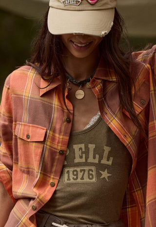 a-woman-wearing-a-rust-orange-and-yellow-plaid-flannel-shirt-over-an-olive-green-graphic-tank-top-paired-with-a-beige-baseball-cap