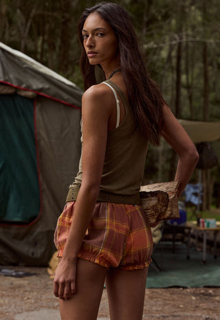 a-woman-standing-outdoors-in-a-campsite-setting-wearing-a-sleeveless-olive-brown-top-and-rust-orange-plaid-shorts-with-a-tent-and-trees-in-the-background