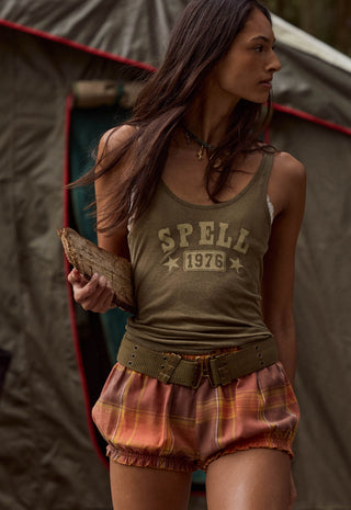 a-woman-wearing-a-vintage-style-olive-green-tank-top-with-spell-1976-printed-on-the-front-paired-with-rust-orange-plaid-biker-shorts-and-a-matching-belt-standing-in-front-of-a-tent