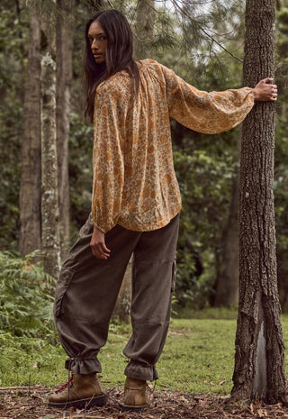 a-woman-stands-in-a-wooded-outdoor-setting-wearing-a-loose-long-sleeve-patterned-top-in-warm-tan-and-brown-tones-dark-olive-cargo-pants-and-brown-hiking-boots-with-one-hand-resting-on-a-tree-trunk