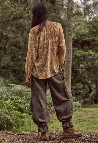 a-person-standing-outdoors-in-a-wooded-area-shown-from-the-back-wearing-a-loose-long-sleeve-blouse-with-an-abstract-print-in-warm-tan-brown-and-cream-tones-paired-with-dark-olive-cargo-pants-and-brown-boots