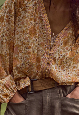 a-close-up-of-a-person-wearing-a-loose-long-sleeve-button-front-blouse-with-a-warm-floral-and-paisley-print-in-tan-rust-and-brown-tones-over-a-cream-base-tucked-into-dark-olive-pants-with-a-brown-belt