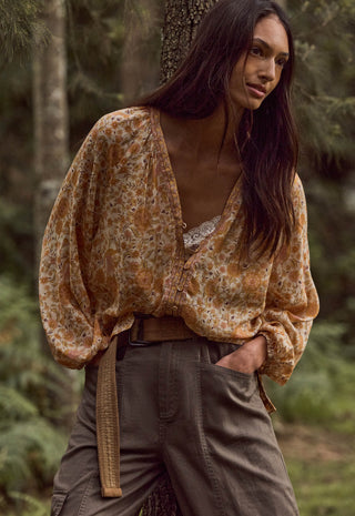 a-woman-standing-outdoors-in-a-forest-wearing-a-loose-long-sleeve-boho-style-blouse-with-an-abstract-floral-pattern-in-tan-brown-and-muted-orange-paired-with-dark-olive-cargo-pants-and-a-woven-belt