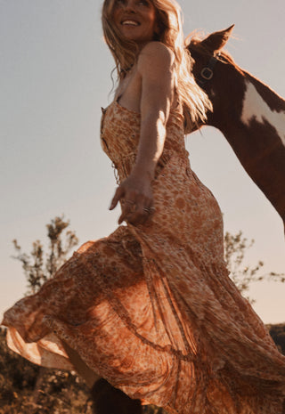 A woman wearing a flowing rust-orange patterned maxi dress is captured outdoors with a brown and white horse beside her against a clear sky.