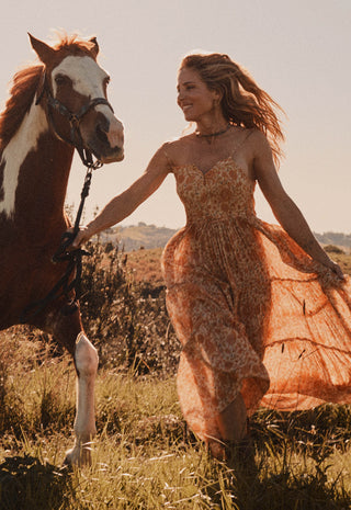 A woman in a flowing rust-orange boho dress walks beside a brown and white horse in an outdoor grassy field at golden hour.