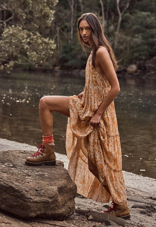A woman stands on a large rock beside a river, wearing a sleeveless, tiered maxi dress in a warm tan and orange floral print, paired with brown lace-up boots.