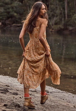 A woman wearing a rust-orange and beige floral print midi dress, standing on a rocky riverbank with water and trees in the background.