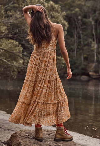 A woman stands on a log by a river wearing a sleeveless, tiered maxi dress with an orange and brown floral pattern. She is shown from behind, looking out over the water with trees in the background.