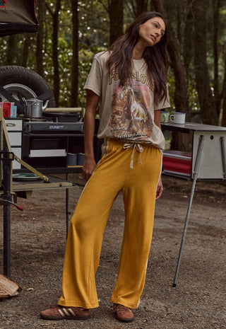 a-woman-stands-outdoors-in-a-campsite-setting-wearing-a-beige-graphic-t-shirt-and-mustard-yellow-wide-leg-sweatpants-she-is-wearing-brown-leather-slide-sandals