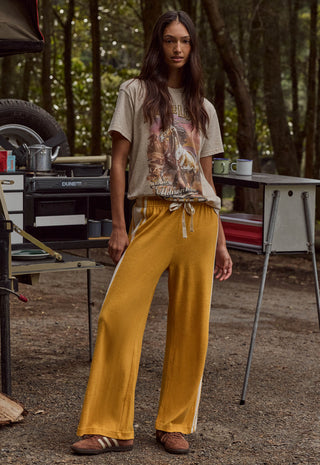 A woman stands outdoors in a wooded campsite setting wearing loose mustard yellow drawstring pants with white side stripes