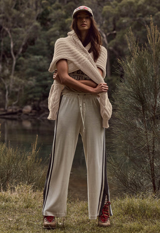 a-woman-stands-outdoors-in-a-wooded-setting-wearing-a-beige-knit-wrap-beige-wide-leg-pants-with-dark-side-stripes-a-baseball-cap-and-red-sneakers