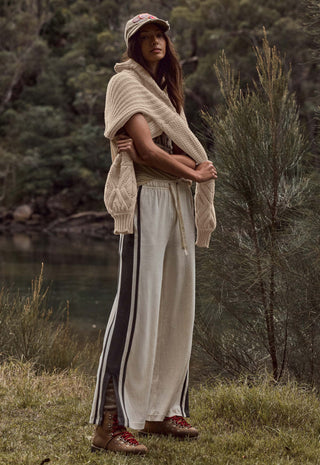 a-woman-stands-outdoors-near-a-body-of-water-and-trees-wearing-a-chunky-knit-beige-wrap-or-poncho-over-cream-wide-leg-pants-with-dark-side-stripes-a-beige-cap-and-brown-lace-up-boots