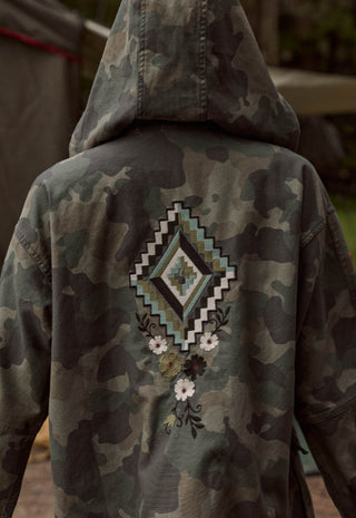 back-view-of-a-camouflage-hooded-jacket-with-a-large-geometric-and-floral-embroidered-design-on-the-back-shown-on-a-person-outdoors