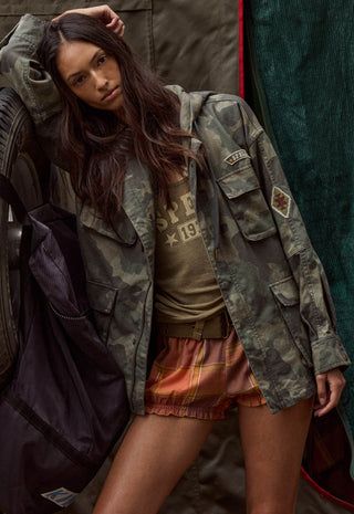 a-woman-posing-in-a-vintage-style-camouflage-utility-jacket-over-a-graphic-t-shirt-and-orange-plaid-shorts-leaning-against-a-rustic-background-with-a-dark-backpack