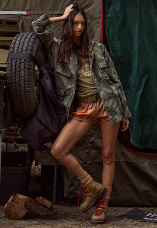 a-woman-poses-in-a-rustic-outdoor-camping-setting-wearing-a-camouflage-utility-jacket-a-graphic-t-shirt-orange-shorts-and-tan-lace-up-boots-with-a-large-spare-tire-and-tent-visible-in-the-background