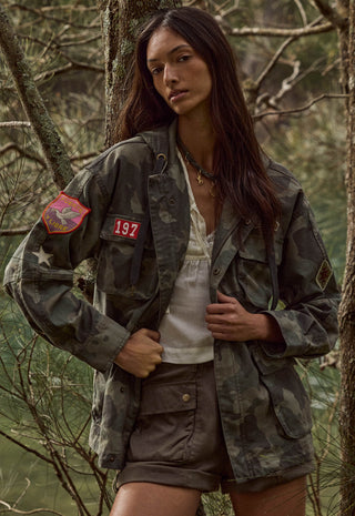 a-woman-wearing-a-military-style-camouflage-jacket-with-red-patches-a-white-top-and-brown-shorts-standing-in-a-wooded-outdoor-setting