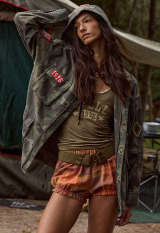 a-woman-wearing-a-vintage-style-olive-green-camouflage-jacket-with-a-red-1976-patch-an-olive-green-graphic-tank-top-and-orange-brown-plaid-shorts-posed-in-front-of-a-rustic-outdoor-tent