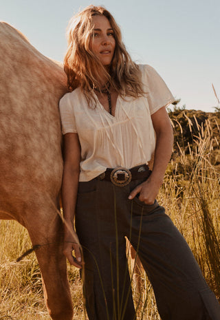 a-woman-stands-outdoors-in-a-field-next-to-a-horse-wearing-a-cream-short-sleeve-blouse-tucked-into-dark-brown-wide-leg-trousers-with-a-western-style-belt-and-large-buckle