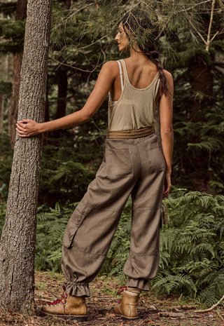 a-woman-stands-in-a-forest-wearing-a-sleeveless-beige-tank-top-loose-brown-cargo-pants-and-brown-hiking-boots-with-her-back-turned-and-one-hand-resting-on-a-tree-trunk