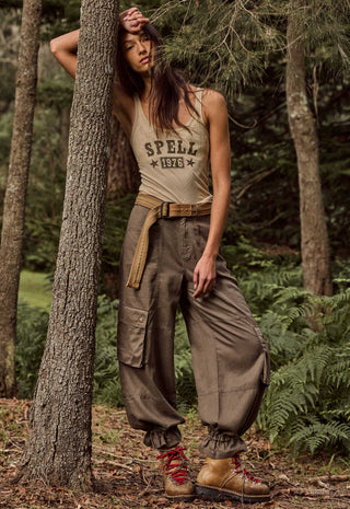 a-woman-posing-in-a-forest-setting-wearing-a-beige-tank-top-with-spell-1976-printed-on-the-front-baggy-brown-cargo-pants-a-tan-belt-and-brown-hiking-boots