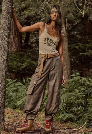 a-woman-stands-in-a-forest-wearing-a-beige-tank-top-with-spells-1976-printed-on-it-loose-brown-cargo-pants-with-a-matching-belt-and-brown-hiking-boots-with-red-laces