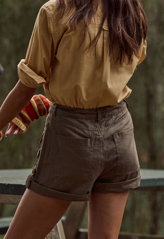 a-person-shown-from-the-back-wearing-olive-brown-utility-shorts-with-rolled-cuffs-and-a-tan-button-up-shirt-standing-outdoors-against-a-blurred-forest-background